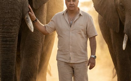 رجلاً عضليًا يمشي حافي القدمين على درب ترابي في السافانا، تحيط به فيلان أفريقيان شاهقان ومهيبان يسيران بمحاذاته